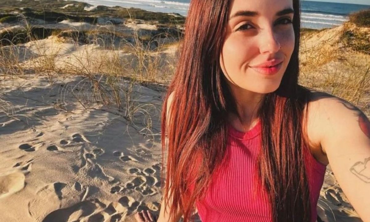 Foto mostra Karyn Lima Souza e Silva, de 24 anos, jovem que desapareceu após pegar uma corrida por aplicativo em SC. Ela está sendo investigada por envolvimento em desvio de R$ 40 mil. Jovem é branca, tem cabelo comprido, veste um cropped rosa e está sentada na areia da praia. Ela posa para selfie