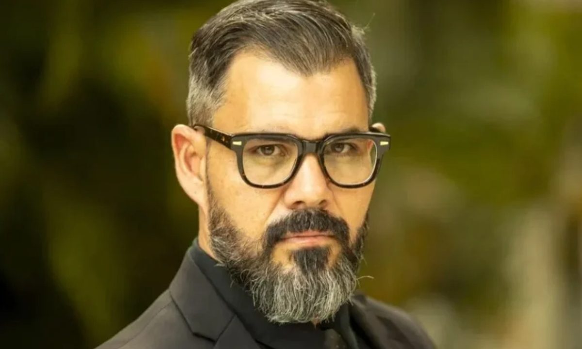 Foto do ator Juliano Cazarré. Ele tem cabelo e barba grisalhos, usa óculos de grau com armação quadrada e escura.