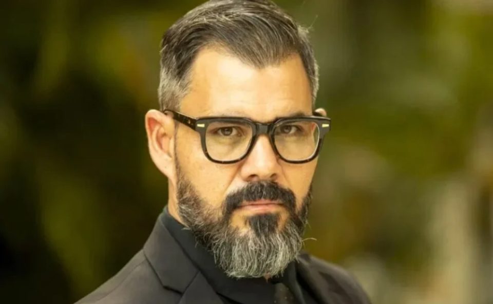 Foto do ator Juliano Cazarré. Ele tem cabelo e barba grisalhos, usa óculos de grau com armação quadrada e escura.
