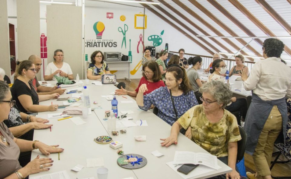 Grupo de pessoas participa de oficina de artesanato em sala do Liceu de Ofícios Criativos, trabalhando em mesas com materiais manuais.