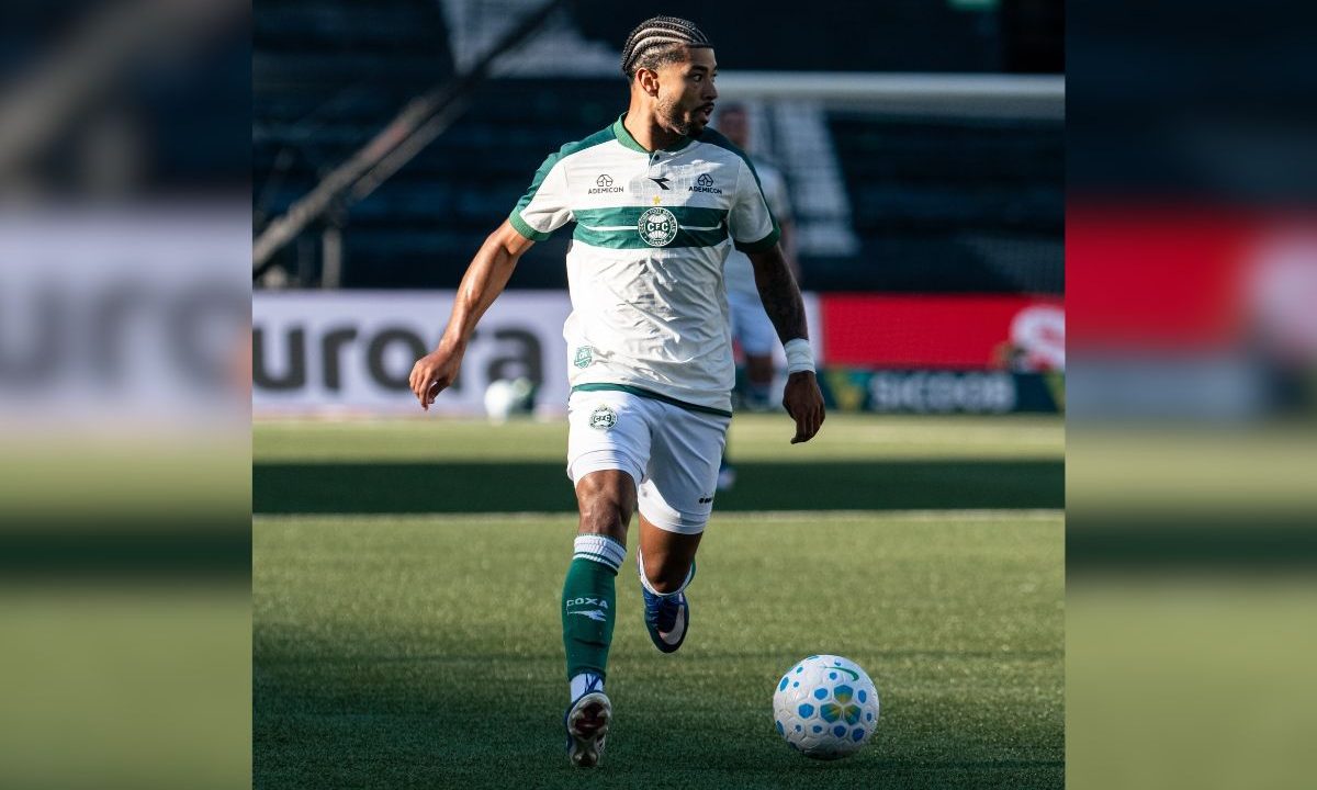 Jogador Lucas Ronier, do Coritiba, conduz a bola em campo durante partida do Brasileirão, vestindo uniforme branco com faixa verde e meias verdes.