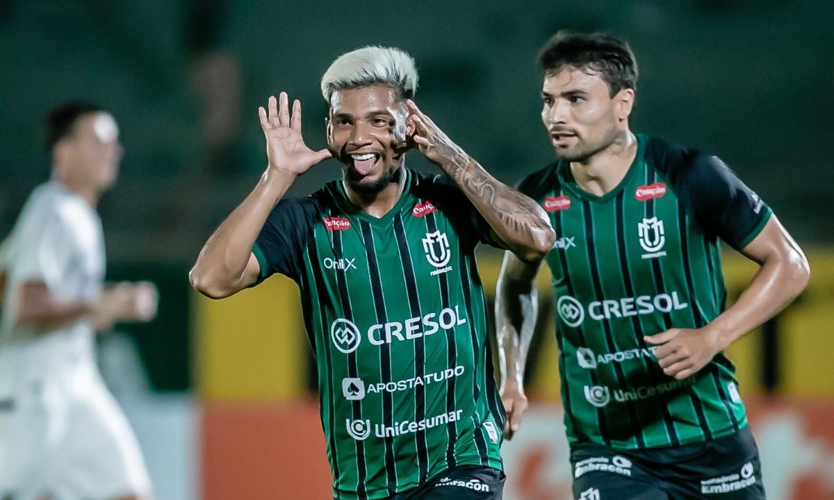 O jogador Negueba, do Maringá, aparece em primeiro plano comemorando um gol com as mãos próximas ao rosto e expressão de alegria. Ele veste uniforme verde com listras pretas. Ao fundo, outro companheiro acompanha a jogada, enquanto o campo aparece desfocado.