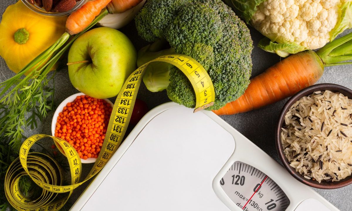 Vista superior de uma balança de banheiro branca cercada por alimentos saudáveis como brócolis, cenoura, maçã verde, lentilhas e arroz. Uma fita métrica amarela está enrolada sobre os alimentos e a balança.