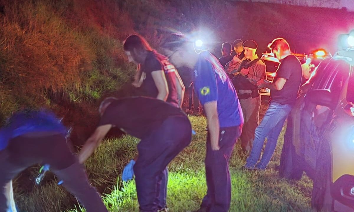Foto mostra policiais e peritos, durante a noite, observando corpo de motociclista que estava desaparecido e foi encontrado morto em uma canaleta da BR-376
