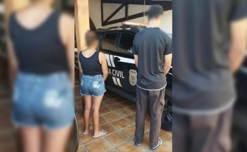 Imagem mostra neta presa por planejar morte da avó com marteladas na cabeça. Ela está ao lado do namorado, executor do crime. Ela usa shorts jeans e regata preta e chinelo. Ele usa calça de moletom marrom, cmaiseta preta. Os dois estão de costas para a foto.
