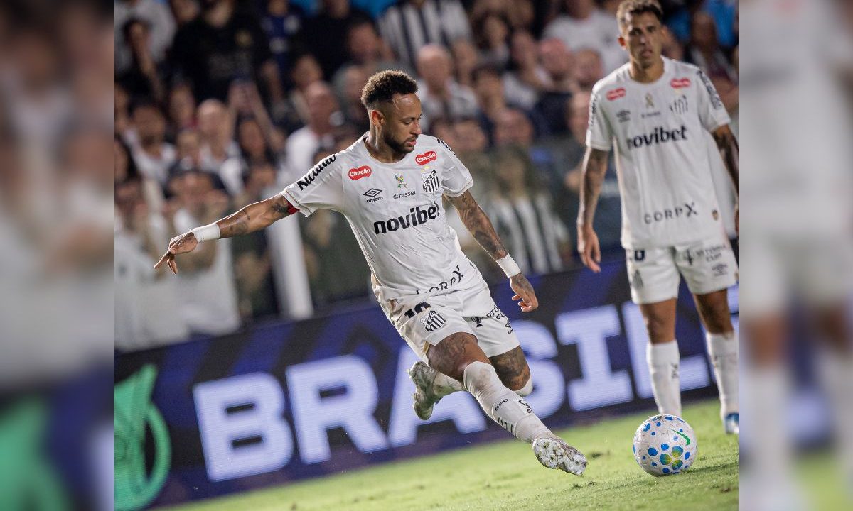 Neymar aparece em campo com o uniforme branco do Santos, no momento em que chuta a bola durante uma partida contra o Remo, na Vila Belmiro. O jogador está levemente inclinado, concentrado na finalização, enquanto um companheiro de equipe observa a jogada ao fundo. A arquibancada está cheia, com torcedores desfocados ao redor do campo. A imagem remete ao debate sobre Neymar na Copa do Mundo e sua presença na Seleção Brasileira
