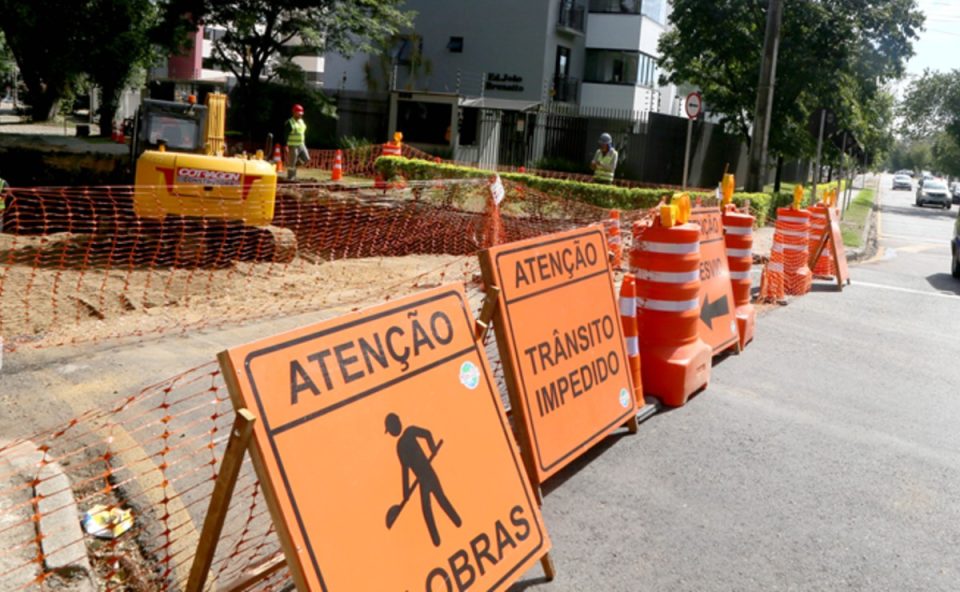 Bloqueio em rua no bairro Portão, em Curitiba, com placas de sinalização indicando obras e interdição. A foto também mostra maquinários e trabalhadores atuando no local.