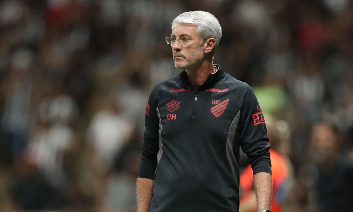O técnico Odair Hellmann aparece à beira do campo, vestindo agasalho preto do Athletico com detalhes em vermelho. Ele observa o jogo com expressão séria, enquanto o fundo mostra a arquibancada desfocada com torcedores.