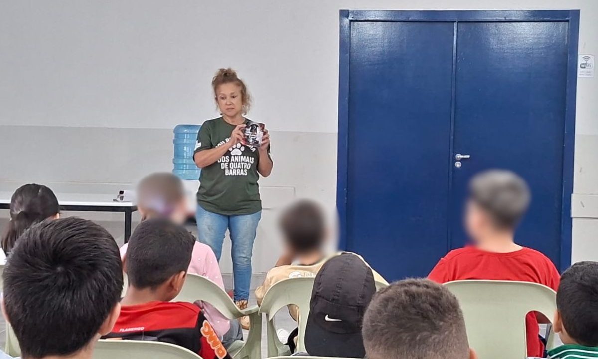 Imagem mostra uma protetora dando palestra sobre maus-tratos aos animais em uma escola para crianças. Ela aparece em pé, com camiseta verde, calça jeans e cabelo preso. As crianças estão sentadas assistindo à palestra.