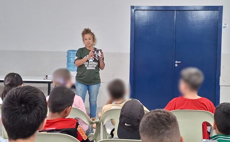 Imagem mostra uma protetora dando palestra sobre maus-tratos aos animais em uma escola para crianças. Ela aparece em pé, com camiseta verde, calça jeans e cabelo preso. As crianças estão sentadas assistindo à palestra.