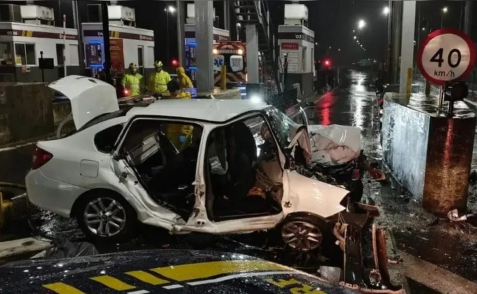 Imagem mostra o carro dirigido por um adolescente completamente destruído em um acidente em uma praça de pedágio em Santa Catarina. O carro é branco e está completamente amassado. Duas mulheres morrem no acidente.
