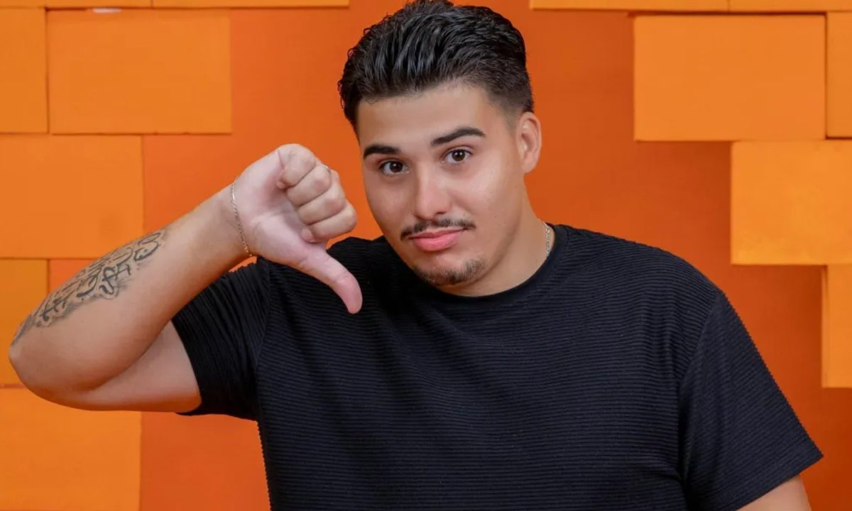 A imagem mostra Pedro Espíndola, ex-participante do BBB 26, que recebeu notificação da Globo. Ele está diante de uma parede laranja com retângulos destacados, fazendo um sinal de negativo.