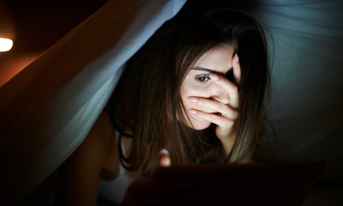 Mulher deitada na cama à noite cobre parte do rosto com a mão enquanto olha para a tela do celular, com expressão de medo e insônia por pesadelos frequentes