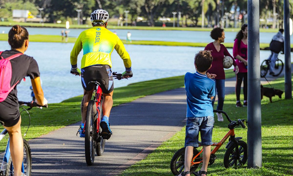 Foto ilustrativa da previsão do tempo para Sexta-Feira Santa em Curitiba. Imagem mostra pessoas andando de bicicleta e crianças brincando no Parque Barigui. A criança veste camiseta azul e bermuda jeans. Ciclistas usam roupas coloridas.