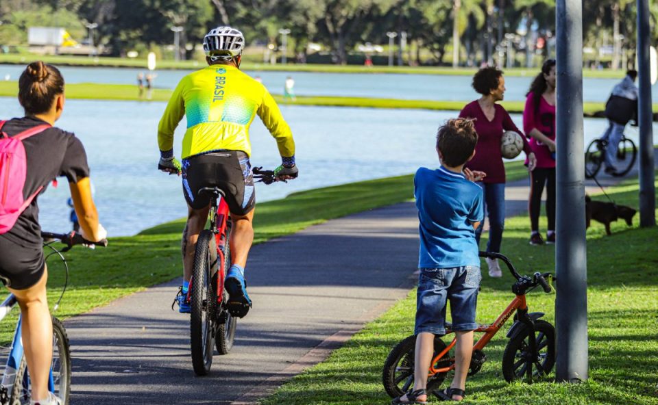 Foto ilustrativa da previsão do tempo para Sexta-Feira Santa em Curitiba. Imagem mostra pessoas andando de bicicleta e crianças brincando no Parque Barigui. A criança veste camiseta azul e bermuda jeans. Ciclistas usam roupas coloridas.