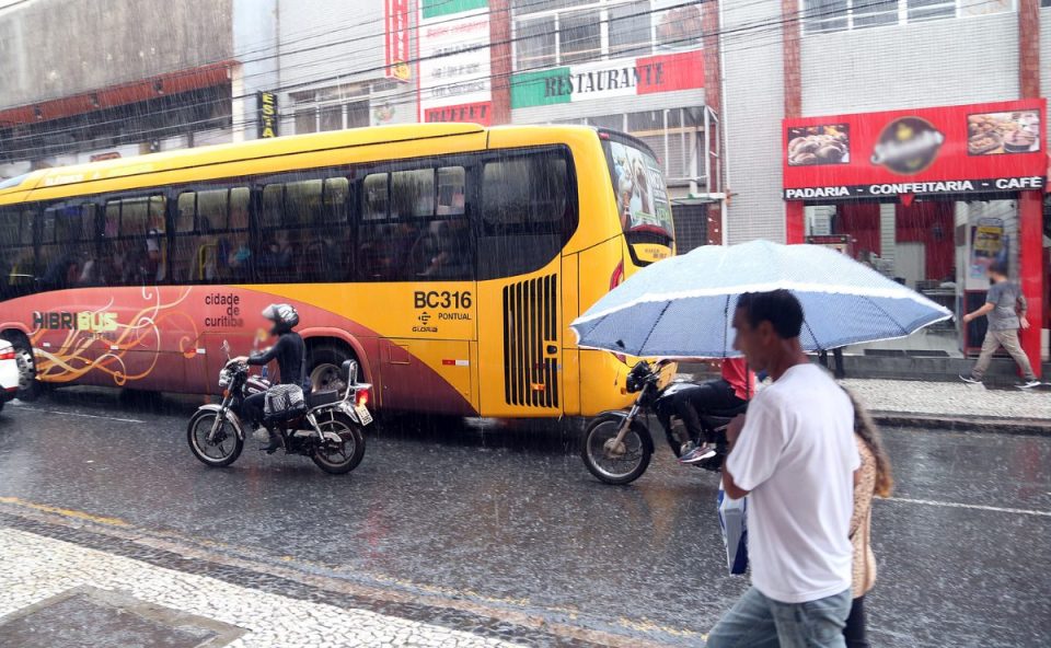 Foto ilustrativa para representar a previsão do tempo em Curitiba nesta terça-feira. Imagem mostra um ônibus ao lado de uma moto. Um homem anda na calçada com um guarda-chuva. O dia está chuvoso. O homem usa camiseta branca e calça jeans.