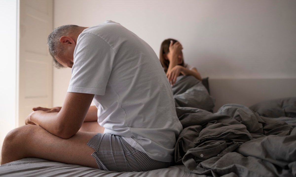 Homem sentado na cama com postura abatida enquanto mulher aparece ao fundo, sugerindo dificuldades no relacionamento íntimo por disfunção erétil, que pode ser tratada com a cirurgia de prótese peniana
