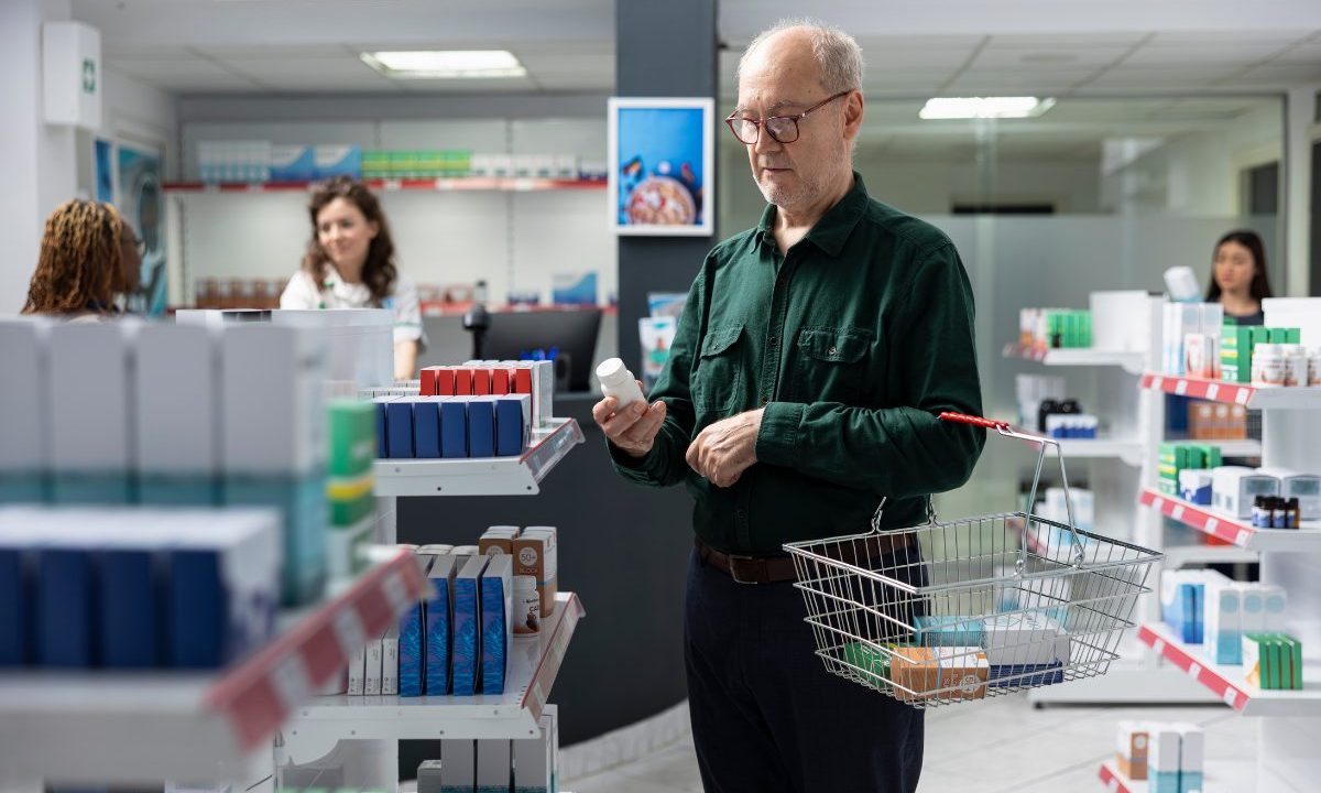 Homem analisa embalagem de medicamento dentro de farmácia instalada em supermercado, segurando uma cesta de compras.