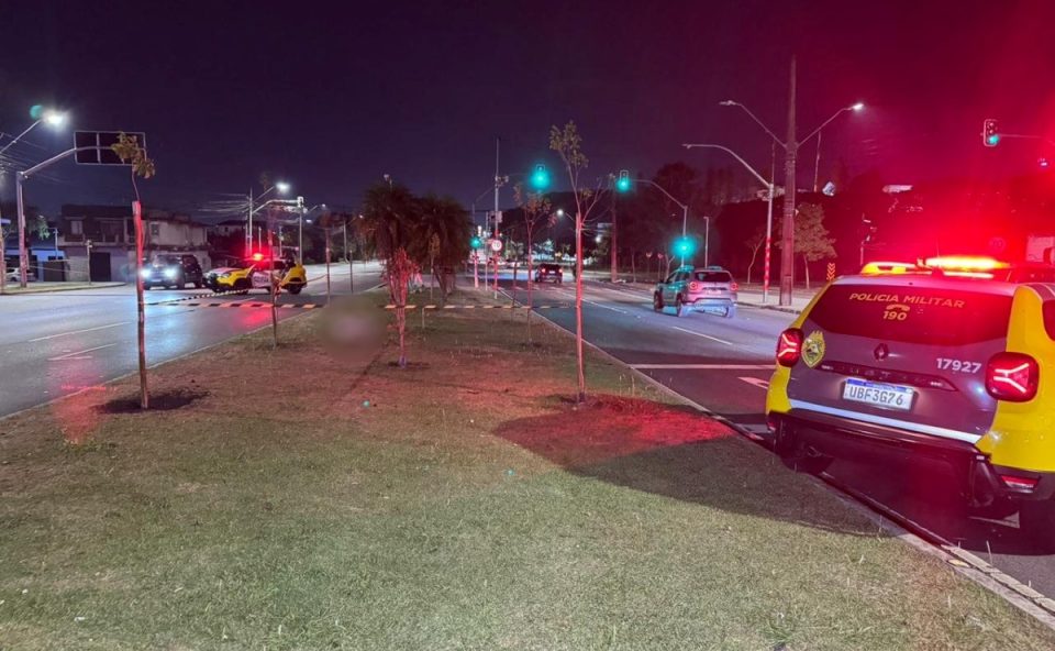 Foto tirada durante a noite mostra local onde suspeito de ferir homem durante assalto foi morto ao atacar policiais em Curitiba. O homem está morto, coberto por uma manta, sobre um gramado. Uma viatura da Polícia Militar aparece próxima ao corpo