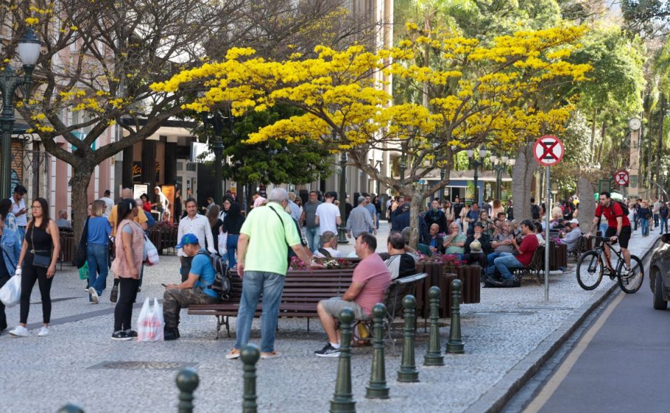 Imagem ilustrativa para a previsão do tempo em Curitiba nesta sexta-feira. Foto mostra diversas pessoas no calçadão perto da Praça da Osório em Curitiba. Tem pessoas sentadas, em pé, em cima de bicicleta, com alto movimento na região. Também há árvores na praça.
