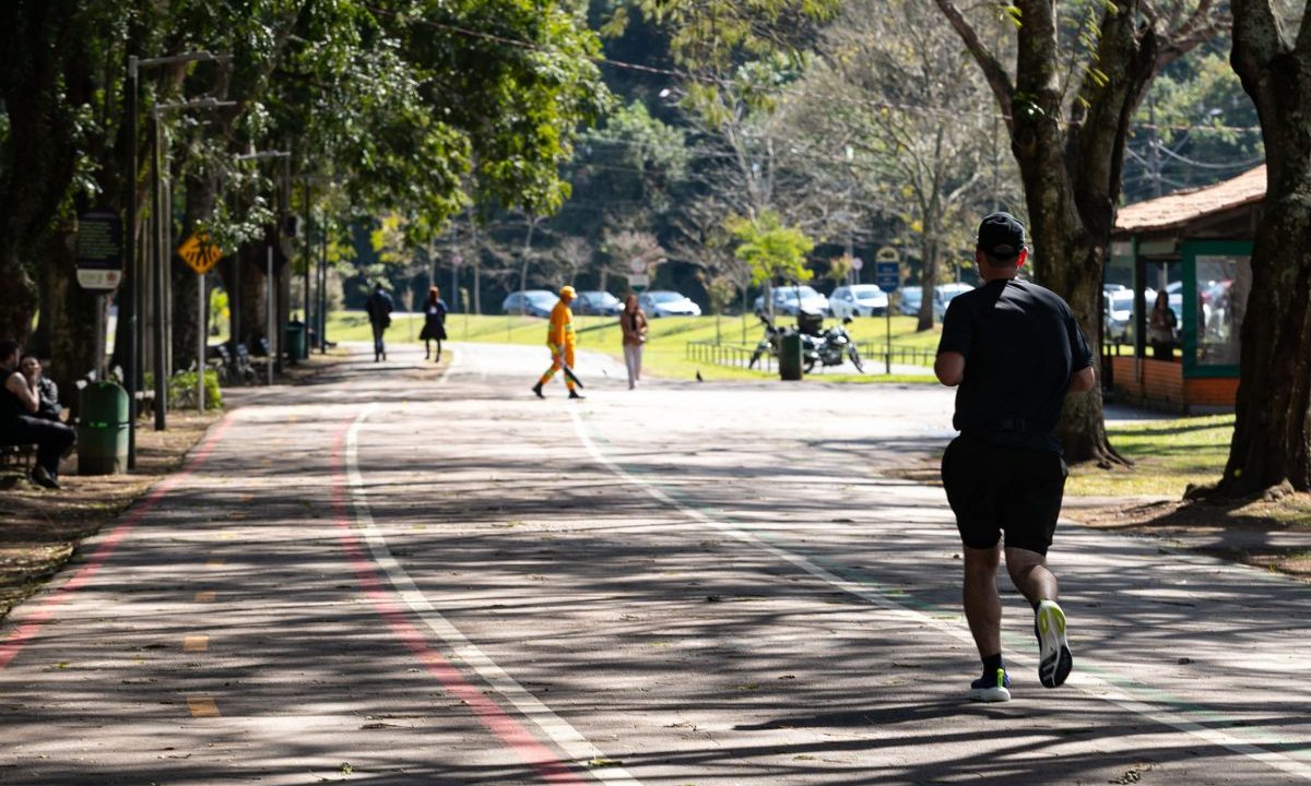 Imagem ilustrativa para previsão do tempo em Curitiba nesta terça-feira. Foto mostra um homem com roupa preta correndo no Parque Barigui. O dia está claro, com pouco sol e bastante sombra de árvores. Outras pessoas também praticam atividades ao ar livre.