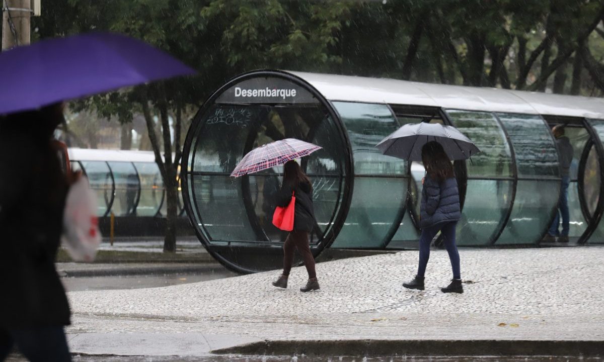 Imagem ilustrativa para a previsão do tempo em Curitiba nesta terça-feira (28). Foto mostra um cenário de chuva, perto de uma estação-tubo. Há pessoas na calçada. Uma mulher com bolsa vermelha e roupa preta segurando guarda-chuva. Outra pessoa está caminhando com jaqueta preta e guarda-chuva também.