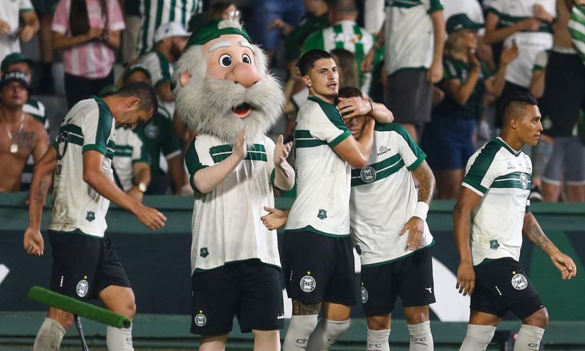 Jogadores do Coritiba em campo no Couto Pereira após lance ofensivo, com Tiago Cóser ao centro abraçando um companheiro, enquanto o mascote do clube aparece ao lado e a torcida alviverde acompanha ao fundo.