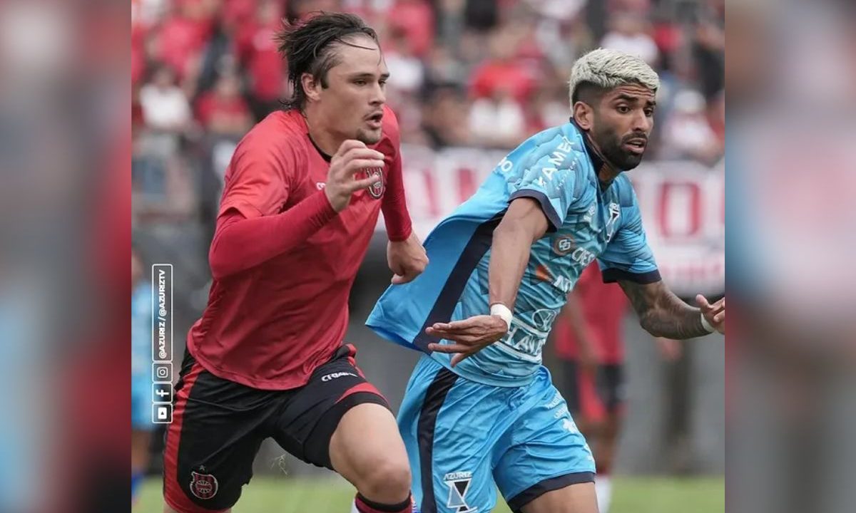Dois jogadores disputam a bola em velocidade, um com uniforme azul do Azuriz e outro com uniforme vermelho do Brasil de Pelotas, durante partida da Série D.