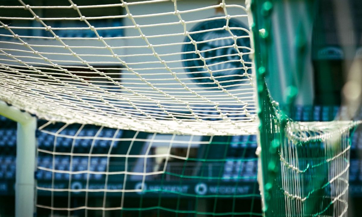 Imagem mostra um close da rede de uma das traves do Couto Pereira, com o fundo desfocado das arquibancadas ao redor do campo. A foto destaca o ambiente de jogo no estádio do Coritiba durante a partida contra o Fluminense.