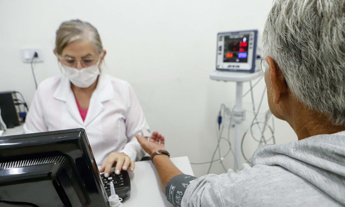 Profissional de saúde mede a pressão arterial de um idoso em consultório. Vacina da gripe e Covid-19 previne complicações como AVC