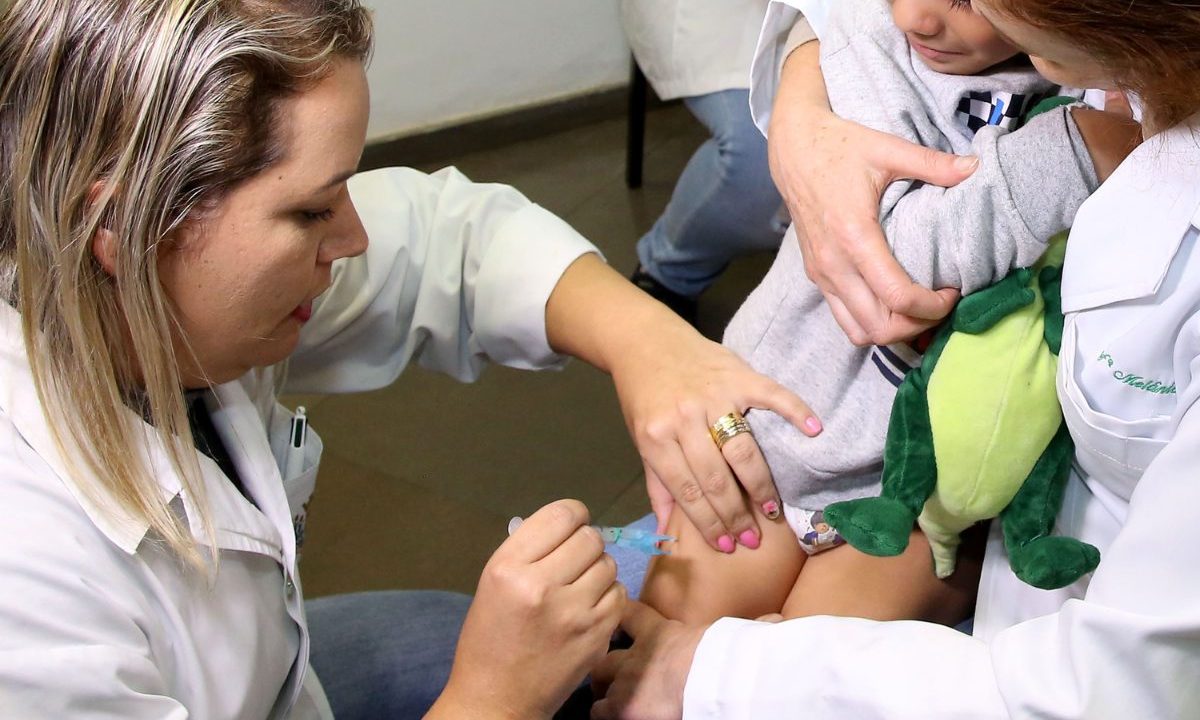 Profissional de saúde aplica vacina em criança no colo da mãe durante atendimento em unidade de saúde.