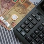 Cédulas de Real (50 e 10 reais), uma moeda de um Real e uma calculadora sobre um gráfico de barras. A imagem ilustra as novas regras do programa Novo Desenrola Brasil para trabalhadores informais e bons pagadores.