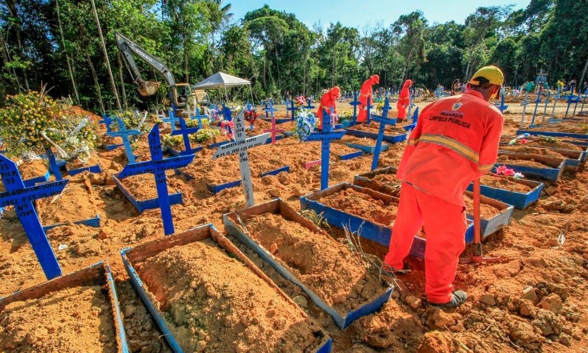 Coveiros com uniformes laranjas trabalham em valas comuns abertas no cemitério durante a pandemia. Fileiras de cruzes azuis marcam as sepulturas recentes em solo de terra avermelhada, cercadas pela floresta.