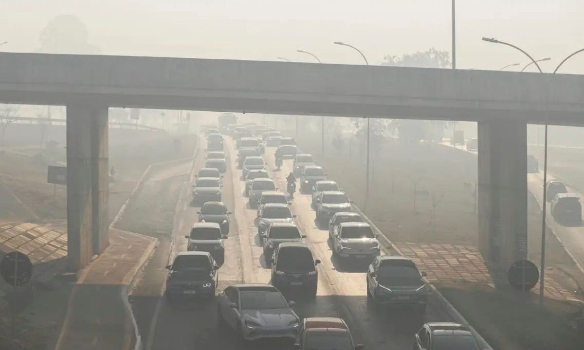 Trânsito intenso de carros em uma rodovia coberta por uma espessa camada de fumaça e poluição sob um viaduto.