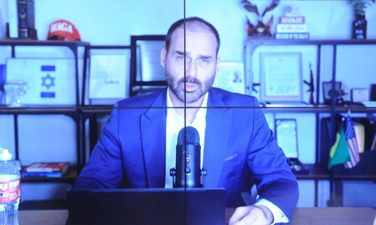 Fotografia de uma tela de monitor dividida por linhas de grade mostrando o ex-deputado Eduardo Bolsonaro. Ele está sentado em frente a um microfone de podcast, vestindo paletó azul e camisa branca. Ao fundo, prateleiras com um boné vermelho escrito "MAGA", uma bandeira de Israel e uma bandeira dos Estados Unidos.