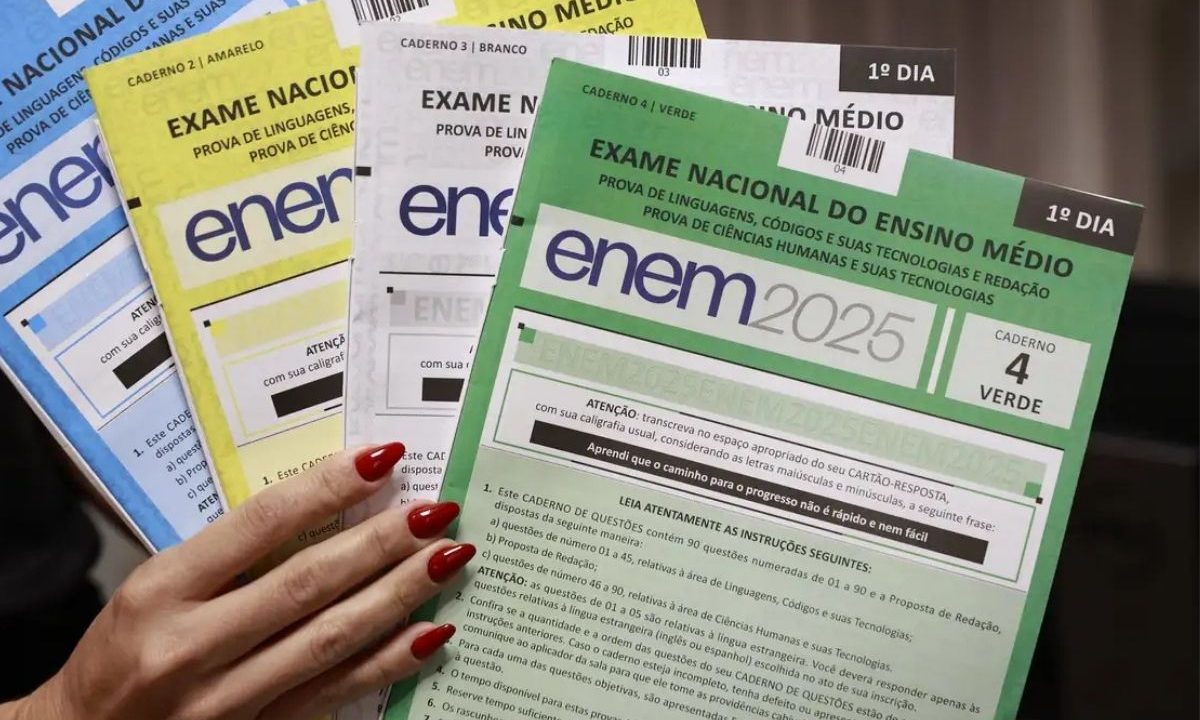 Mão segurando quatro cadernos de questões do Enem de cores diferentes (azul, amarelo, branco e verde), ilustrando o resultado dos pedidos de isenção na taxa do Enem 2026.