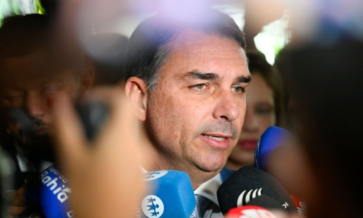 Foto em close-up do senador Flávio Bolsonaro falando com jornalistas, cercado por microfones no Senado.