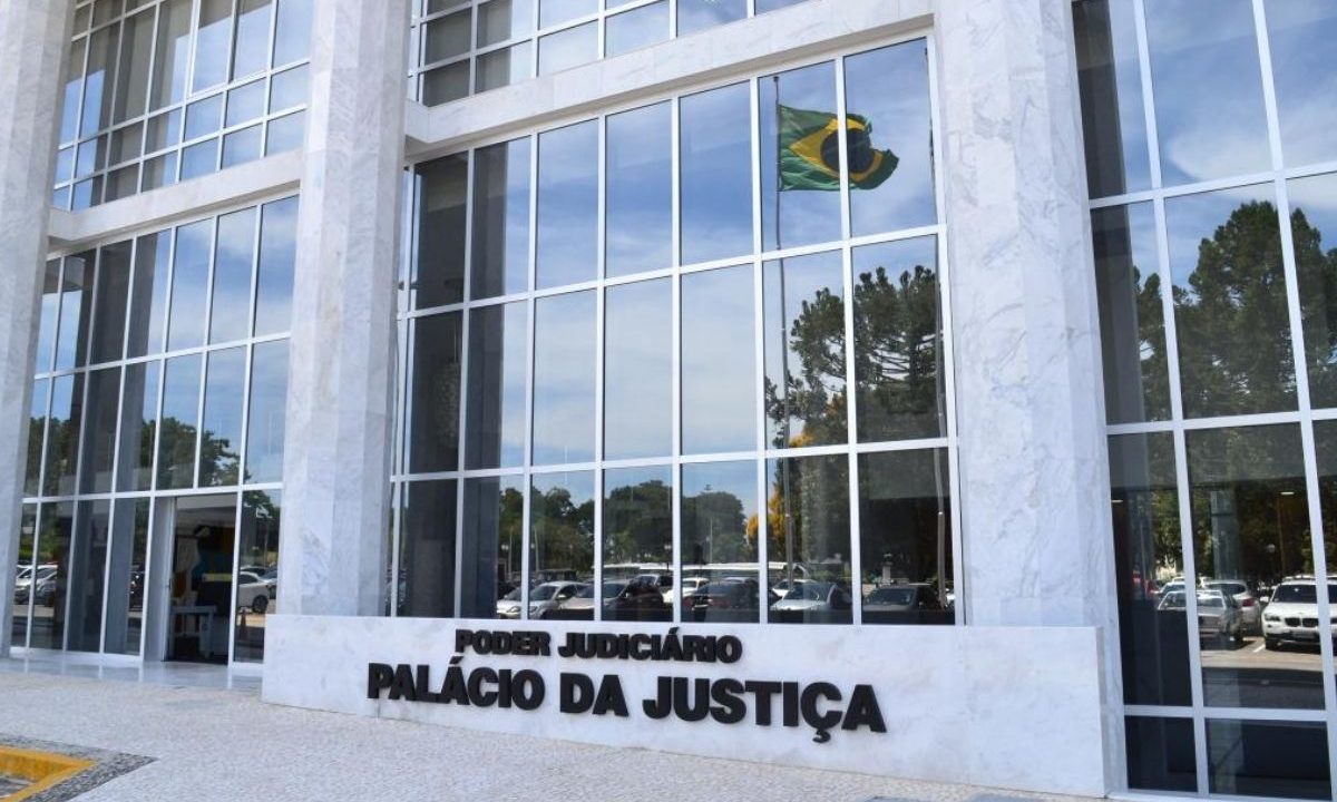 Fachada do Palácio da Justiça, sede do Tribunal de Justiça do Paraná (TJ-PR) em Curitiba, com destaque para a placa de identificação em mármore e o reflexo da bandeira do Brasil nos vidros do edifício.