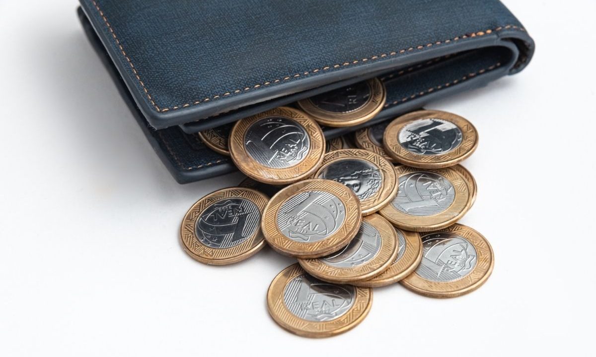 Carteira azul aberta com diversas moedas de 1 Real brasileiro saindo de dentro sobre um fundo branco.