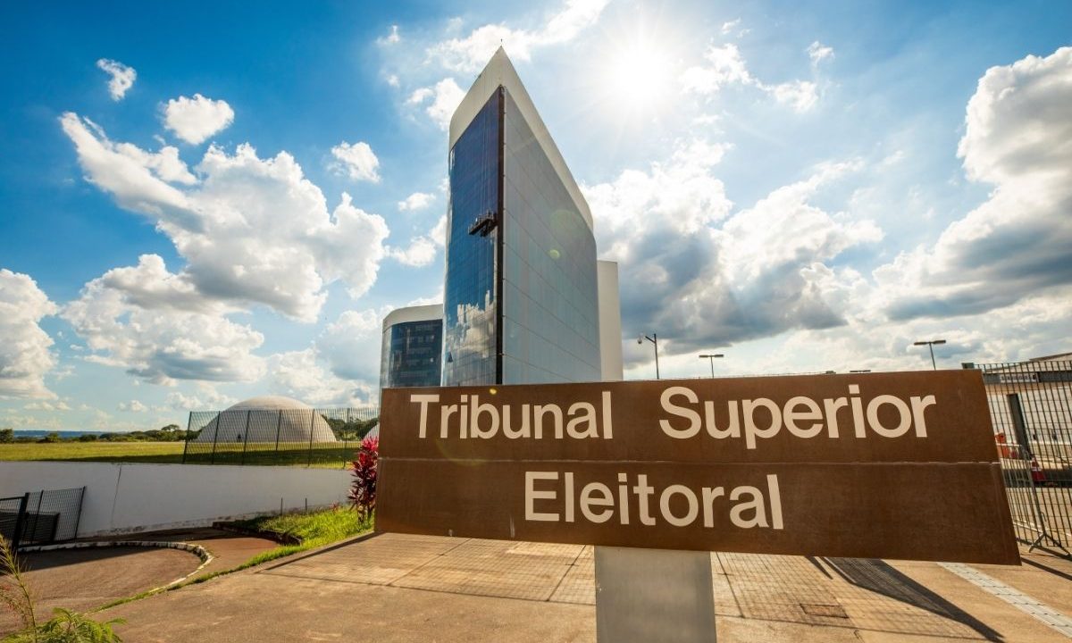 Fachada do edifício sede do Tribunal Superior Eleitoral (TSE) em Brasília, com uma placa marrom em destaque no primeiro plano e o prédio espelhado ao fundo sob um céu azul ensolarado