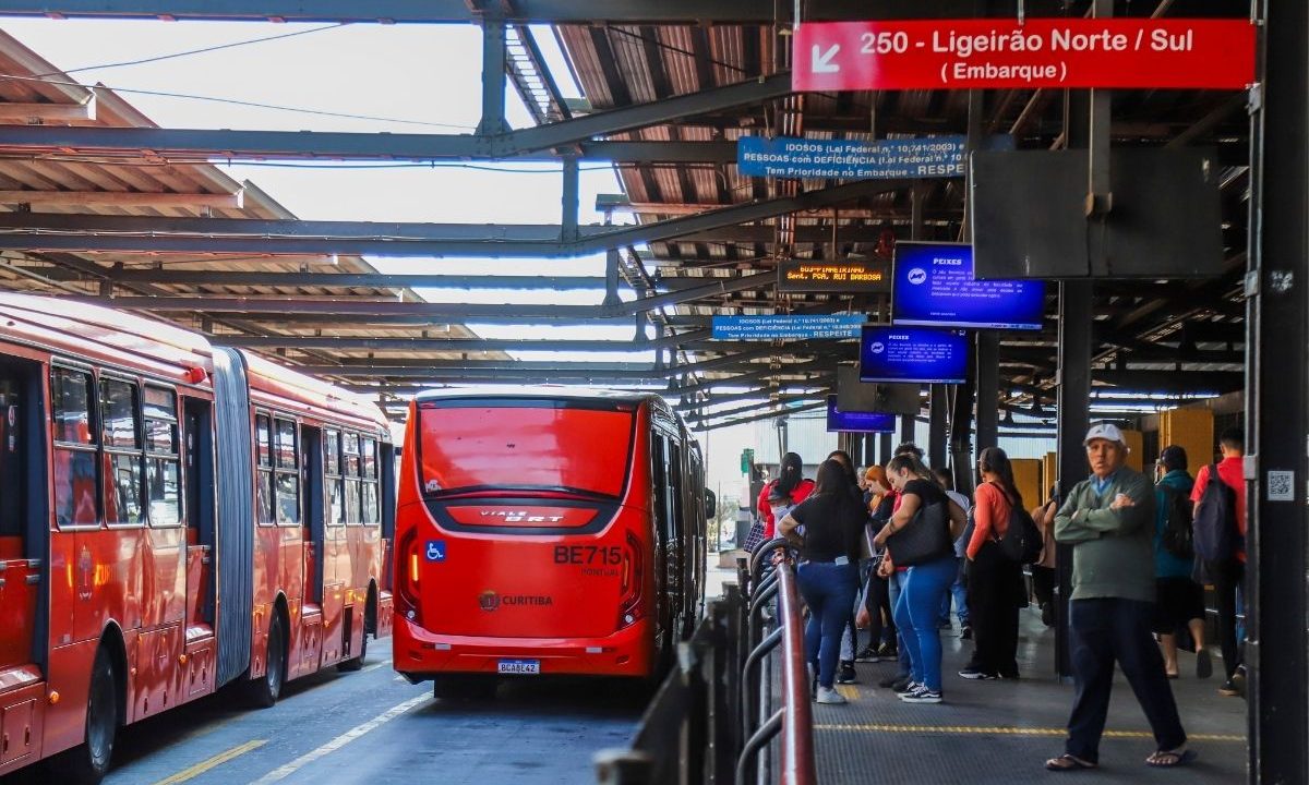 Ônibus biarticulados vermelhos em estação-tubo de Curitiba com passageiros aguardando na plataforma, ilustrando reportagem sobre os impactos do fim da escala 6x1 no transporte coletivo urbano.