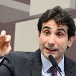 Foto de meio-corpo de Gabriel Galípolo, presidente do Banco Central, falando de forma enfática e gesticulando com a mão direita levantada em forma de garra. Ele está em um pódio com um microfone à frente, vestindo terno cinza, camisa azul clara e gravata listrada. O fundo é um painel acústico geométrico da sala de comissões do Senado Federal.
