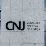 Fachada externa do prédio do Conselho Nacional de Justiça com a sigla CNJ gravada em uma parede de mármore cinza. A foto ilustra as mudanças no sistema que acelera o bloqueio de valores em conta devedores.
