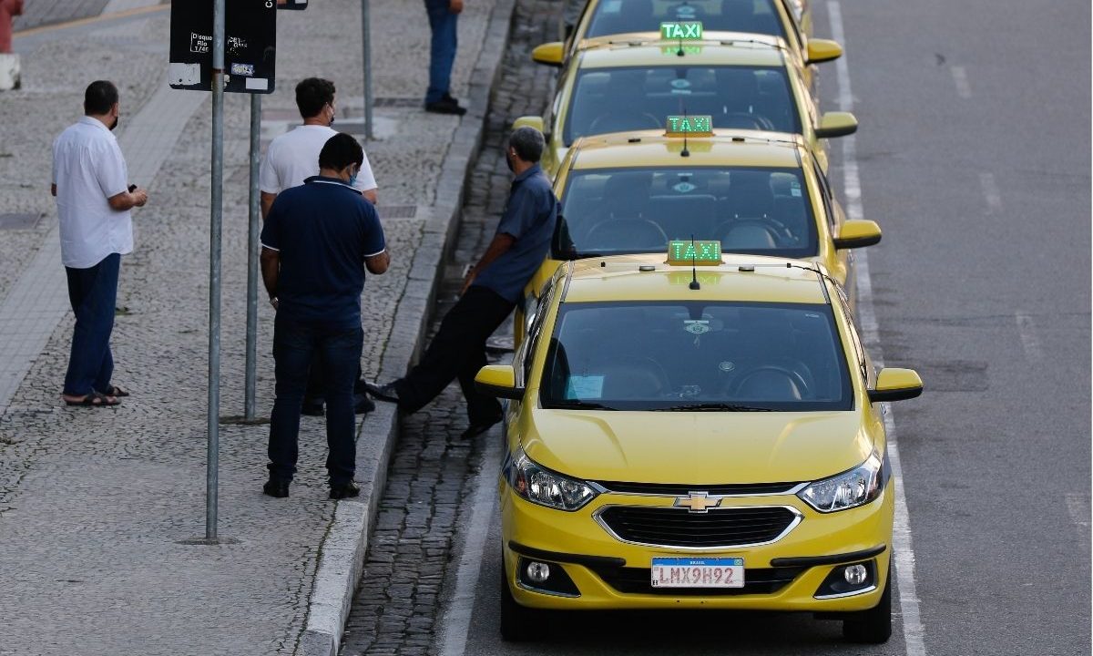 Fileira de táxis amarelos estacionados em um ponto na calçada enquanto os motoristas conversam em pé. A imagem ilustra a aprovação de novas linhas de crédito para taxistas e motoristas de aplicativo.