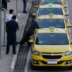 Fileira de táxis amarelos estacionados em um ponto na calçada enquanto os motoristas conversam em pé. A imagem ilustra a aprovação de novas linhas de crédito para taxistas e motoristas de aplicativo.
