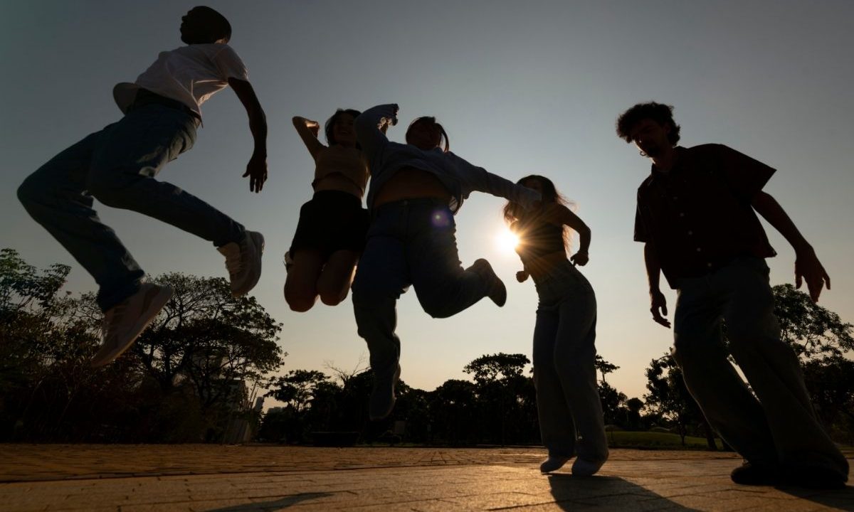 Silhueta de cinco adolescentes pulando e dançando alegremente em um parque ao ar livre durante o pôr do sol. O sol brilha fortemente entre eles, criando um efeito de contraluz. Ao fundo, árvores contornam o horizonte sob um céu claro.
