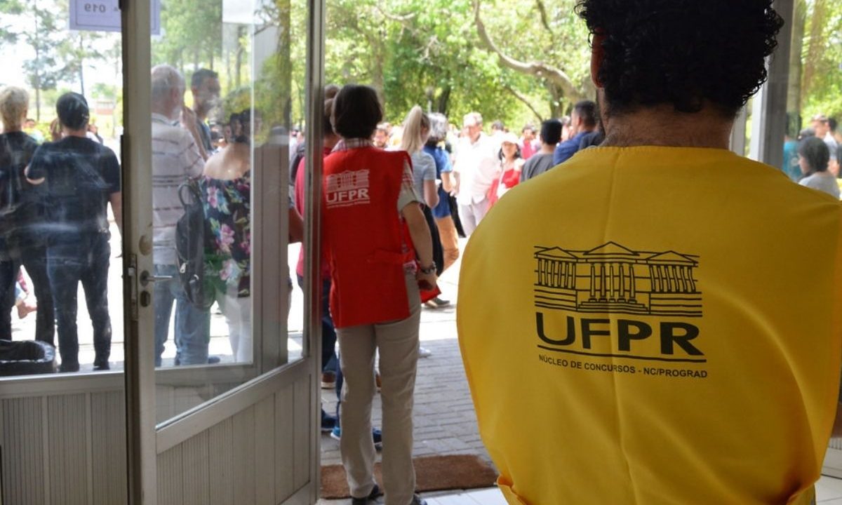 Foto tirada de dentro para fora de um local de prova. No primeiro plano, à direita, aparece de costas um fiscal vestindo um colete amarelo com o logotipo da UFPR e a inscrição "Núcleo de Concursos - NC/PROGRAD". Ao fundo, pelo vão da porta de vidro aberta, vê-se outra fiscal de colete vermelho e um grupo de pessoas aguardando na área externa arborizada.