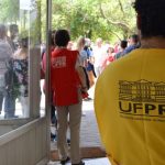 Foto tirada de dentro para fora de um local de prova. No primeiro plano, à direita, aparece de costas um fiscal vestindo um colete amarelo com o logotipo da UFPR e a inscrição "Núcleo de Concursos - NC/PROGRAD". Ao fundo, pelo vão da porta de vidro aberta, vê-se outra fiscal de colete vermelho e um grupo de pessoas aguardando na área externa arborizada.