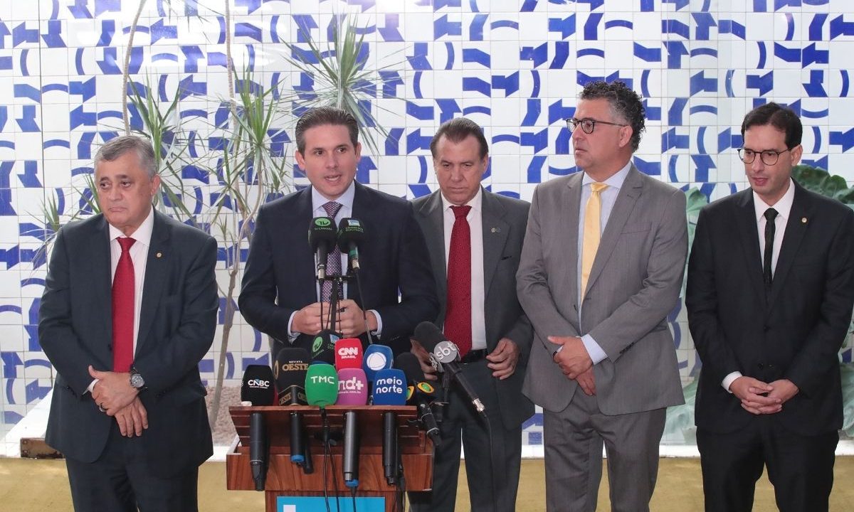 Cinco homens de terno e gravata estão alinhados lado a lado concedendo uma entrevista coletiva em Brasília. O homem ao centro fala ao microfone em um púlpito de madeira repleto de microfones de emissoras de TV e rádio. Ao fundo, há uma parede revestida de azulejos brancos com desenhos geométricos em azul e um vaso com folhagens.