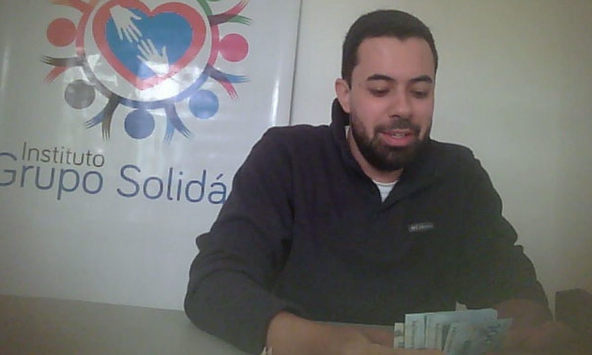 Captura de tela de um vídeo em baixa resolução mostrando o vereador Lórens Nogueira (PP), vestindo um casaco preto. Ele está sentado atrás de uma mesa cinza e segura um maço de cédulas de dinheiro (notas de cem reais) nas mãos. Ao fundo, à esquerda, há um banner com o logotipo de um coração formado por mãos e a inscrição parcial "Instituto Grupo Solidário"